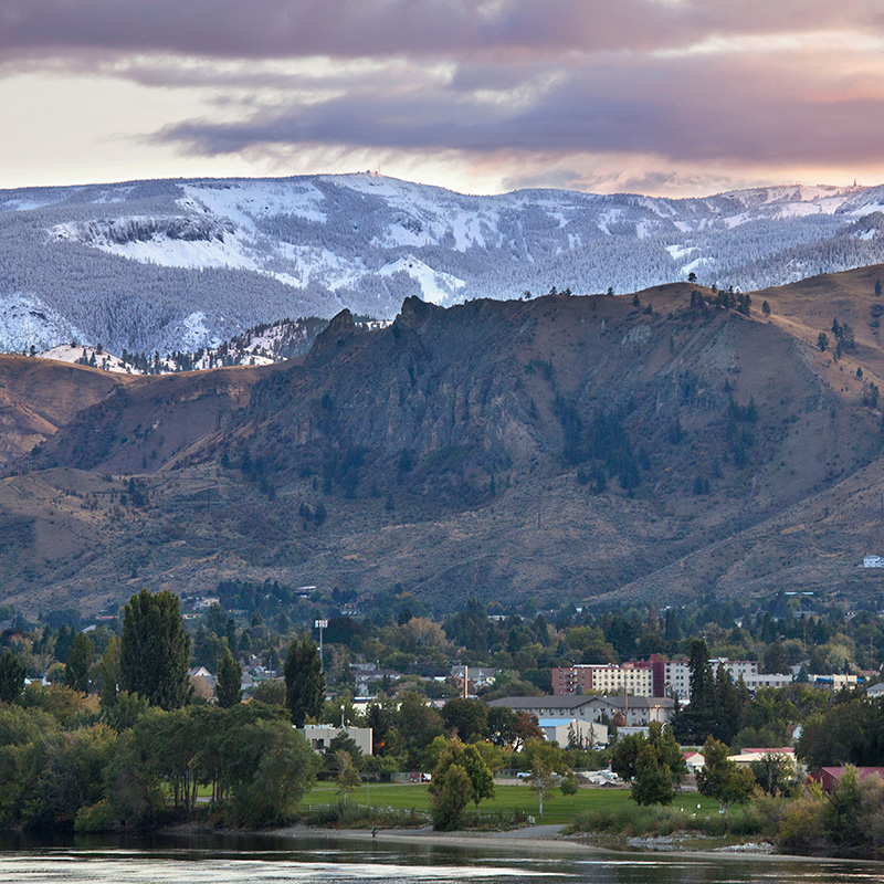 View of a town nestled at the base of a rugged mountain range with early-season snow covering the peaks and forested slopes in the background. The scene is set during a colorful sunset, with dramatic clouds and soft lighting reflecting off a river in the foreground.